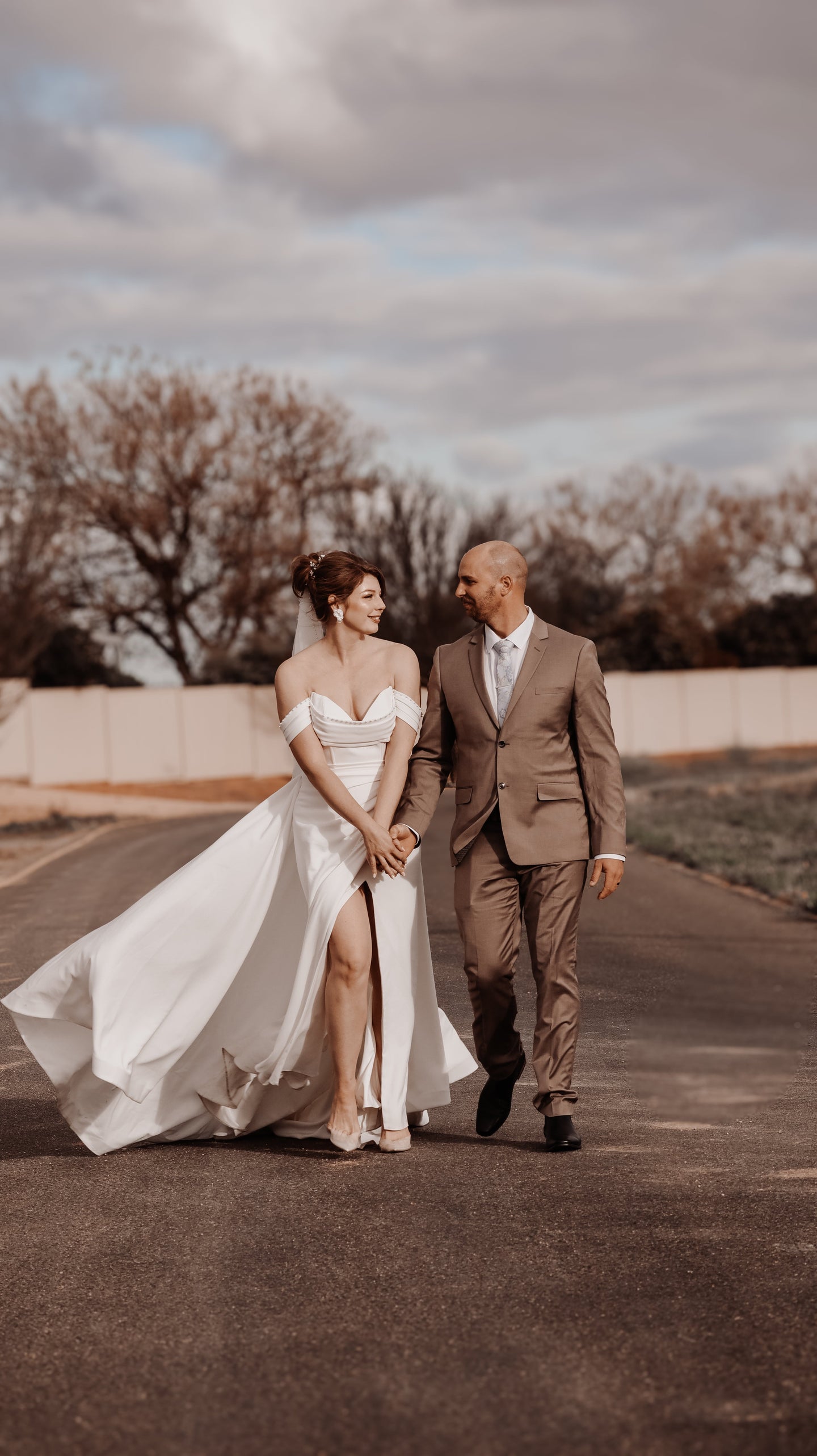 A bride and groom walking along road with a cloudy sky behind them