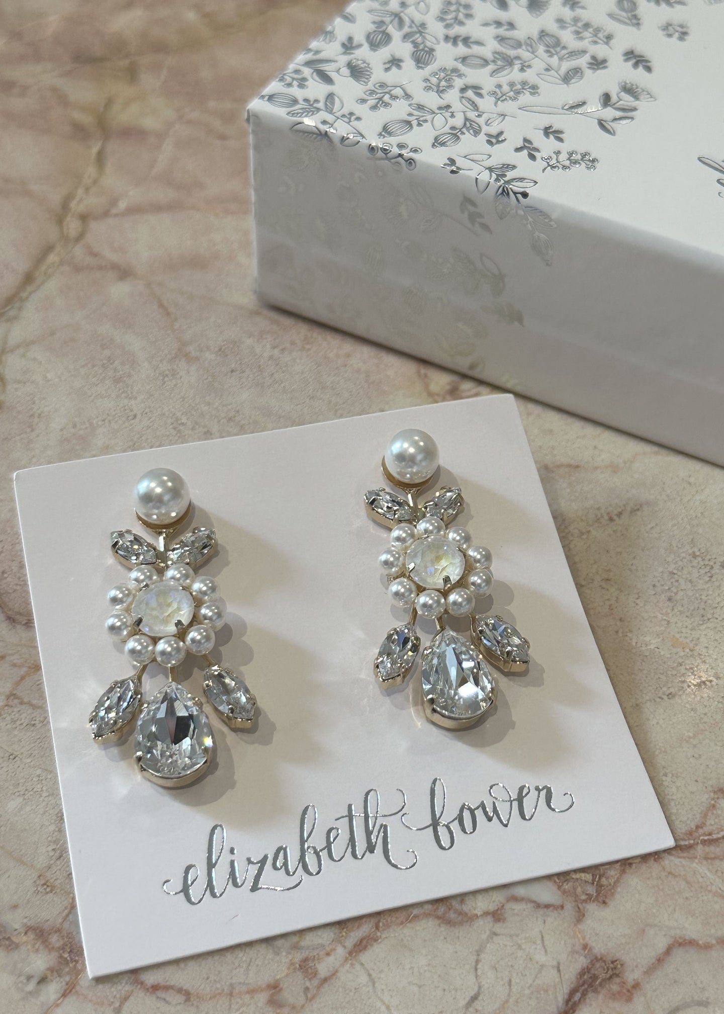 A pair of crystal and pearl drop earrings on an ear card set on a marble background