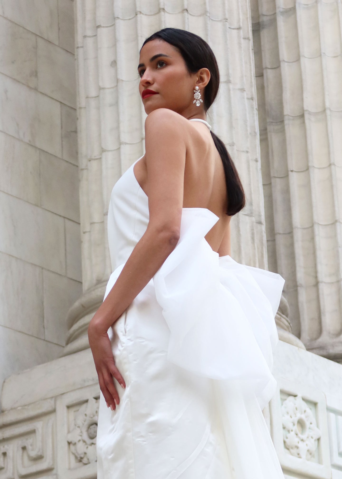 A lady wearing a crystal drop earring and a bow backed wedding dress standing outsite the New York Public Lirbary
