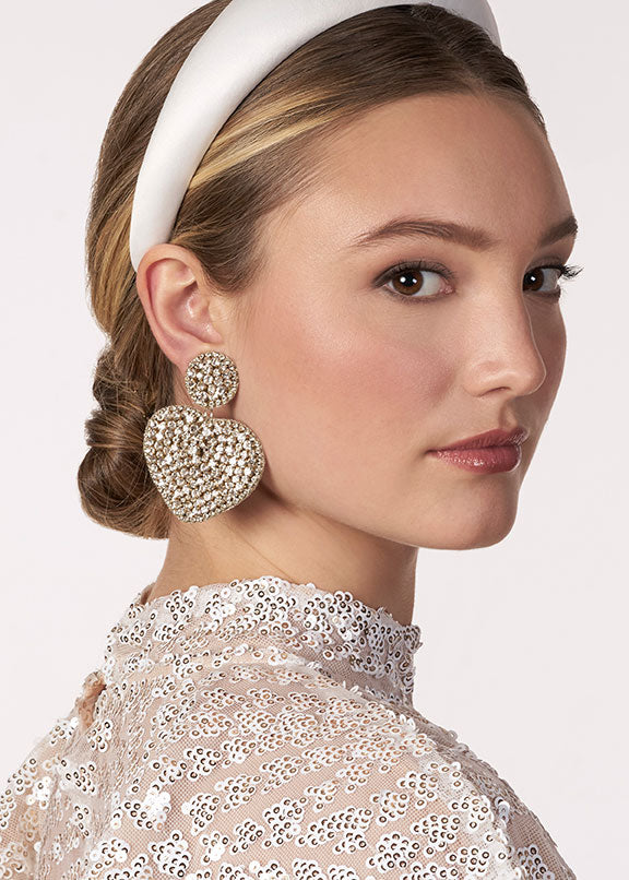 Woman wearing sparkling heart-shaped earrings and a white headband against a neutral background