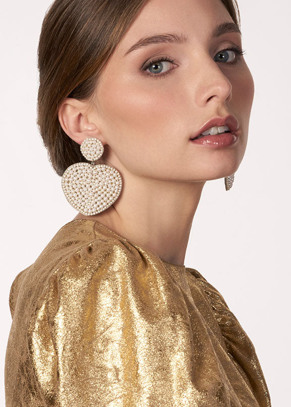 Woman wearing large, round, beaded earrings with a gold topcoat on a plain background