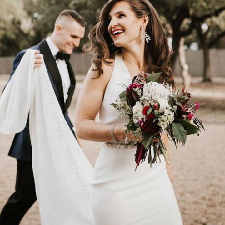 A statement chandelier earring on a smiling brides while the groom carries her dress train