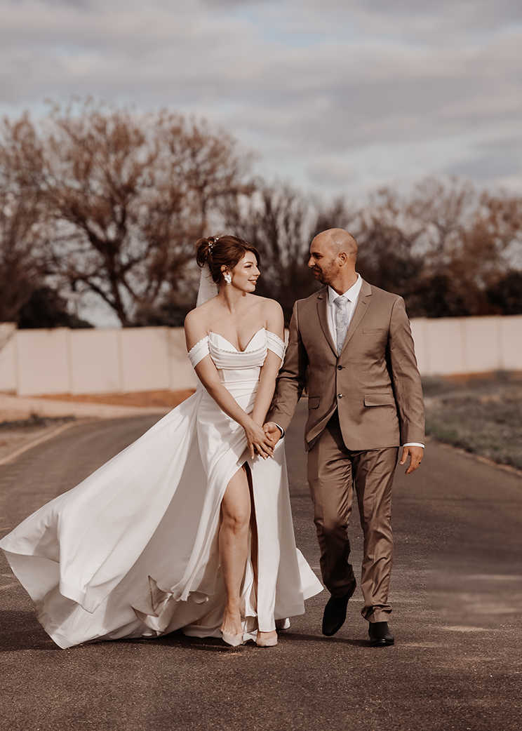 Couple in wedding attire walking outdoors with trees and sky in the background