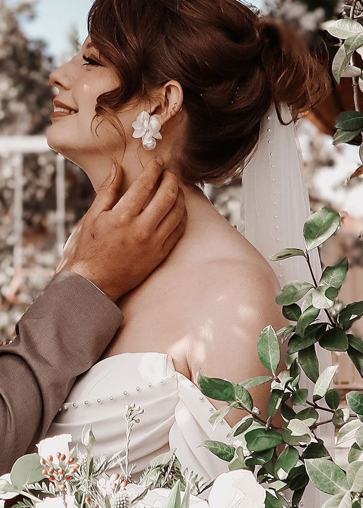 Woman in a wedding dress with floral earrings and a veil, surrounded by greenery.