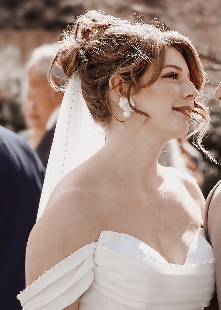Woman in a wedding dress with a veil and pearl earrings smiling outdoors.