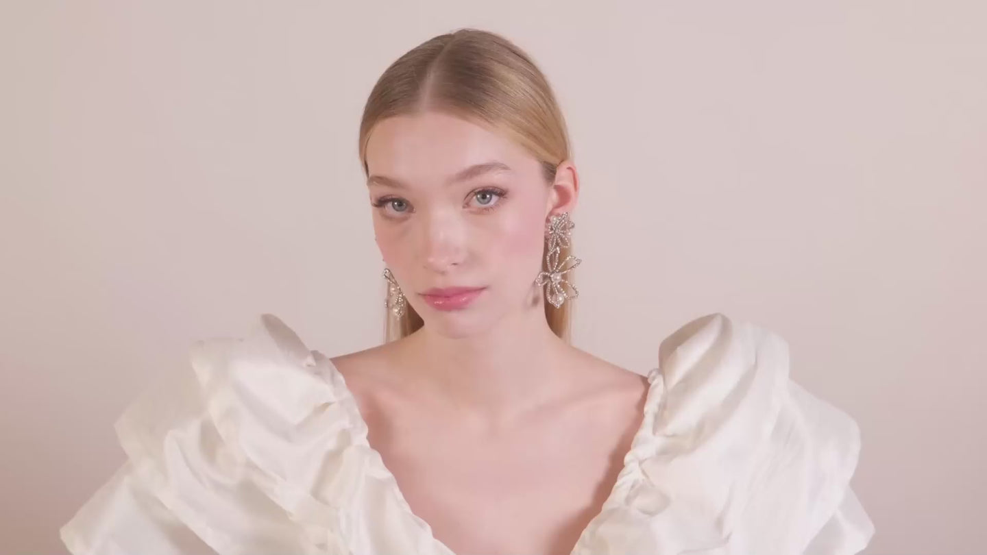 A lady wearing pear and crystal floral earrings in a ruffles silk dress on a beige background